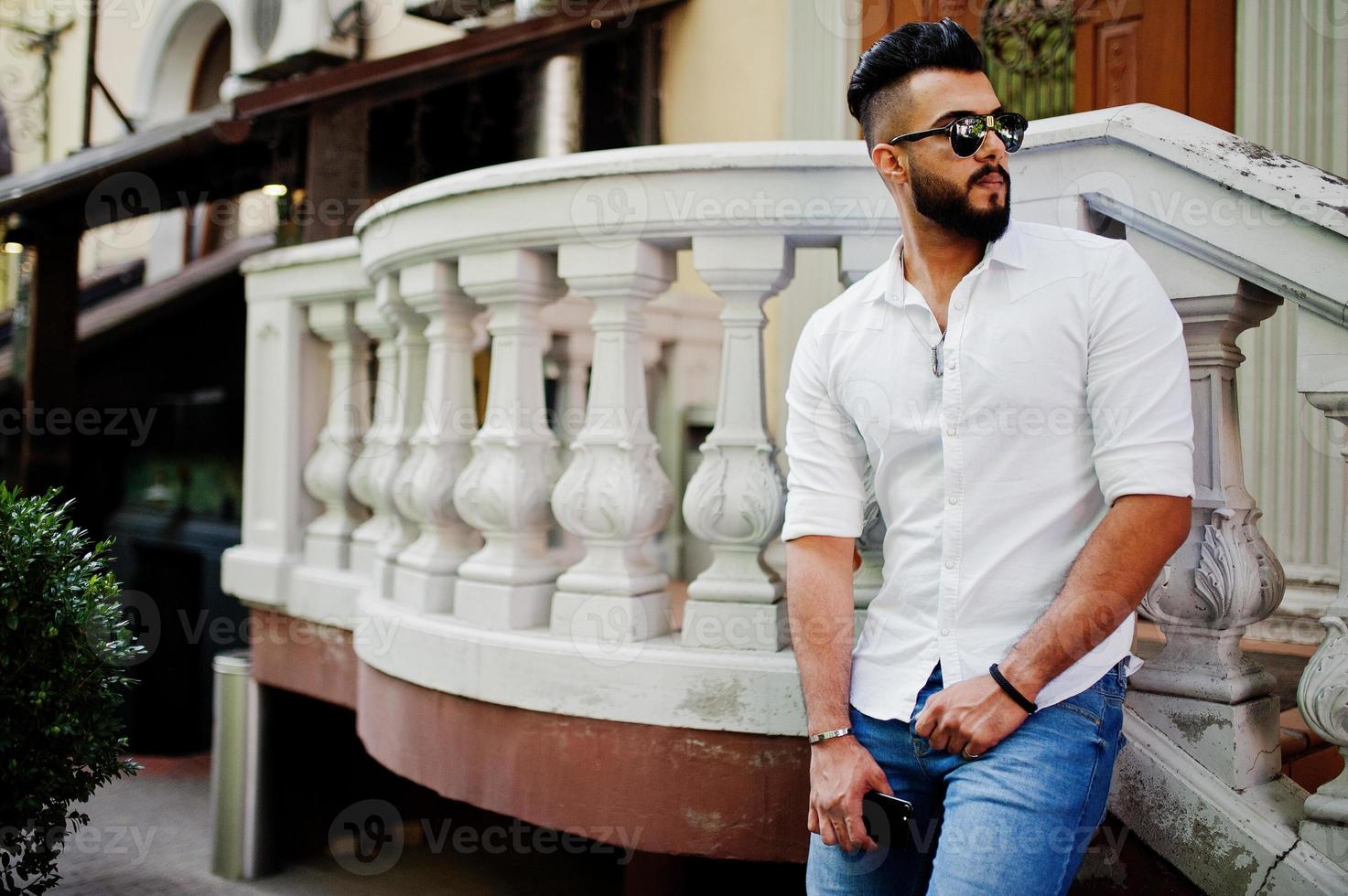 Stilvolles großes arabisches Mannmodell in weißem Hemd, Jeans und Sonnenbrille posierte auf der Straße der Stadt. Bart attraktiver arabischer Typ. foto