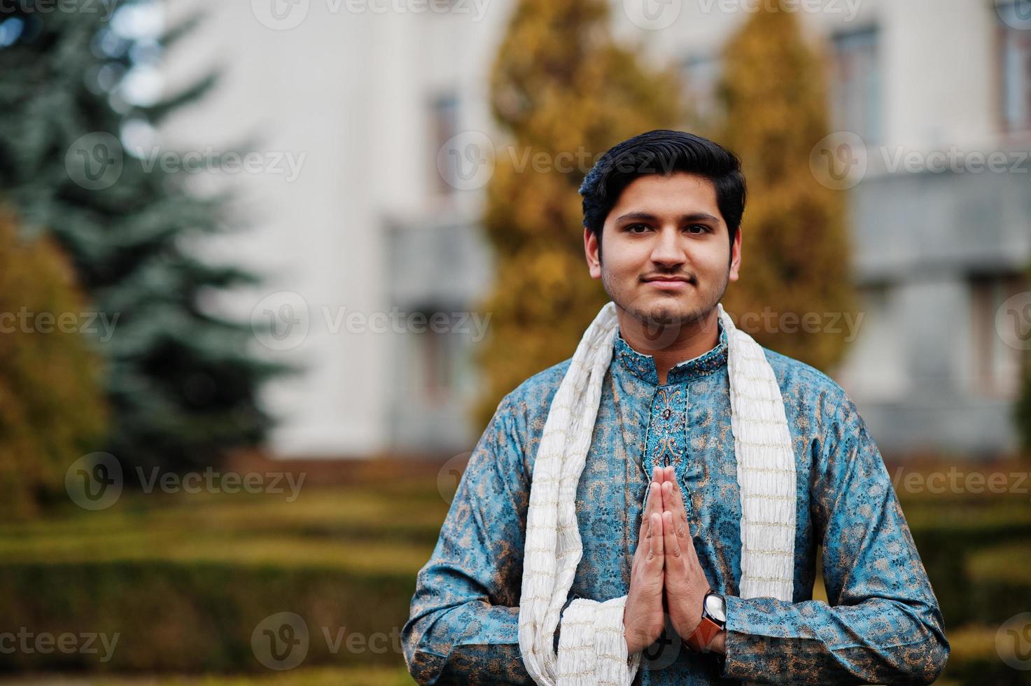 indischer mann trägt traditionelle kleidung mit weißem schal, der im freien gegen grüne büsche im park posiert, zeigt namaste-handzeichen. foto