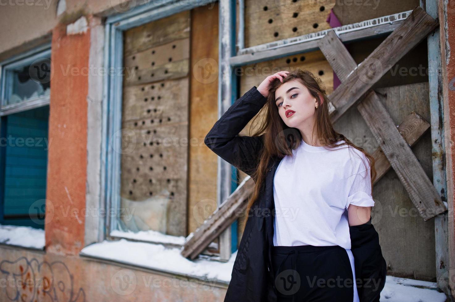 Modisches brünettes Modell mit langen Beinen im langen schwarzen Mantel posierte im Freien am Wintertag gegen alte Grunge-Wand mit zerbrochenen Fenstern. foto