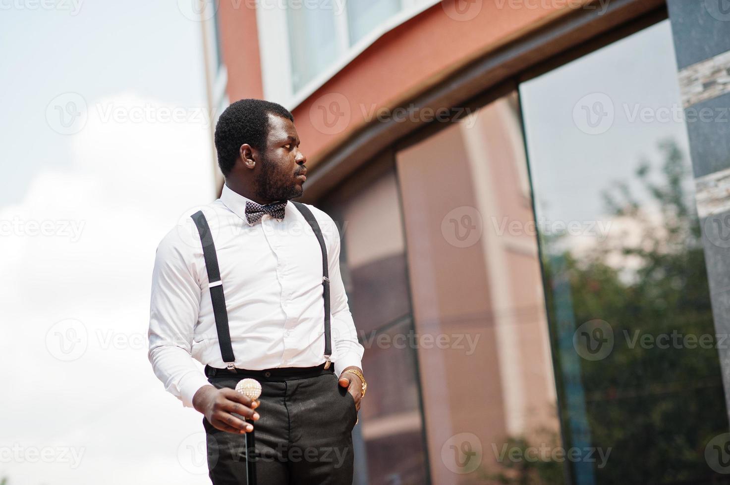 hübscher modischer afroamerikanischer mann in formeller kleidung, fliege und hosenträgern, spazierstock. foto
