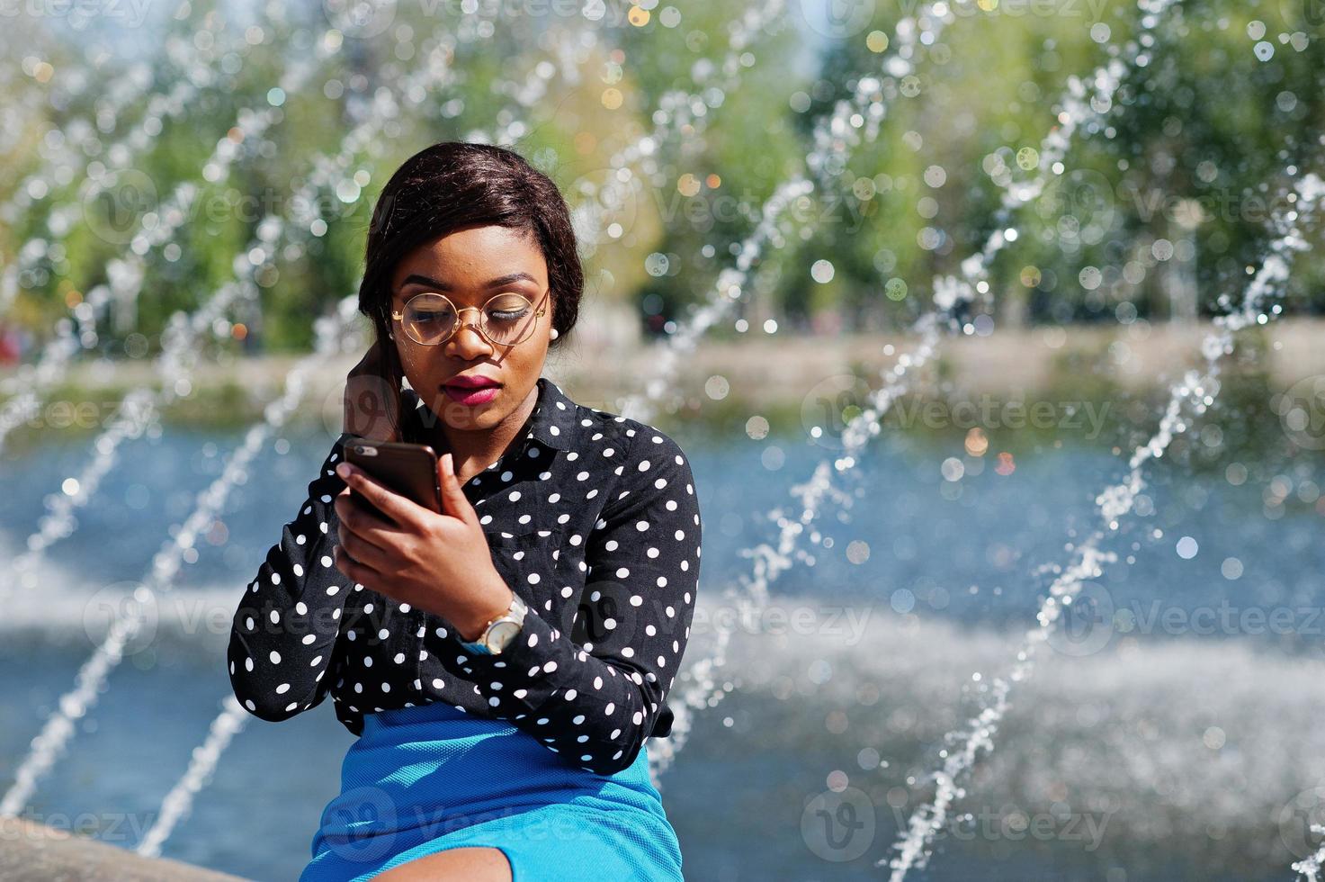 Stilvolles afroamerikanisches Modelmädchen mit Brille, blauem Rock und schwarzer Bluse posierte im Freien mit Springbrunnen und schaute aufs Handy. foto
