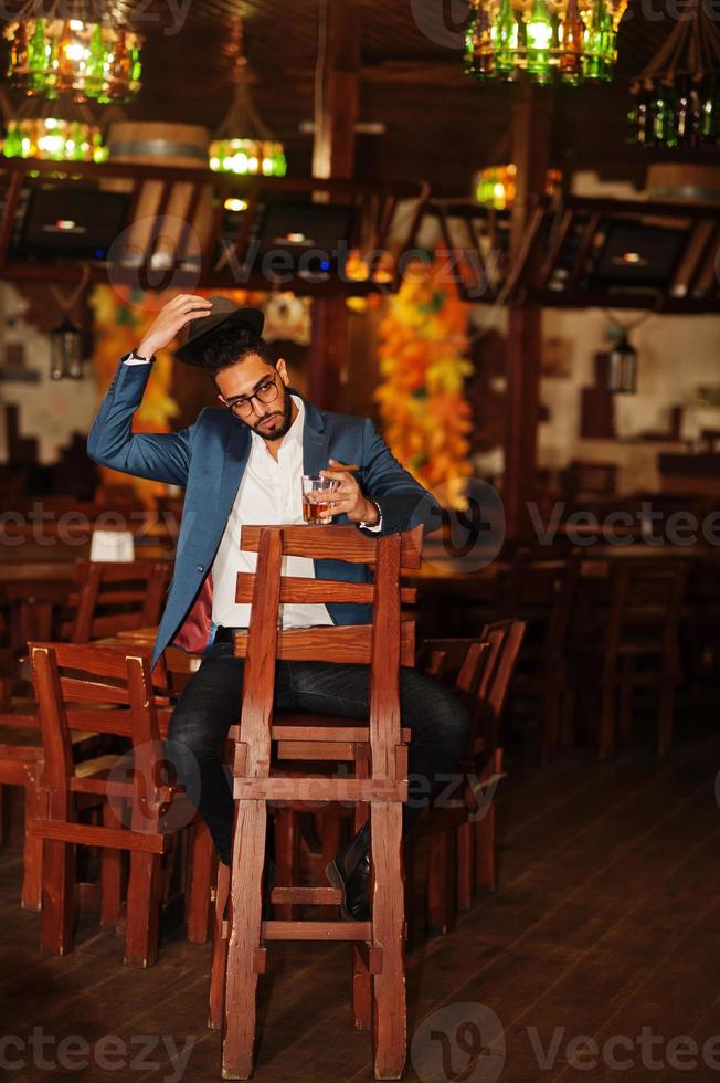 hübscher, gut gekleideter arabischer mann mit einem glas whisky und einer zigarre, die im pub posiert. foto