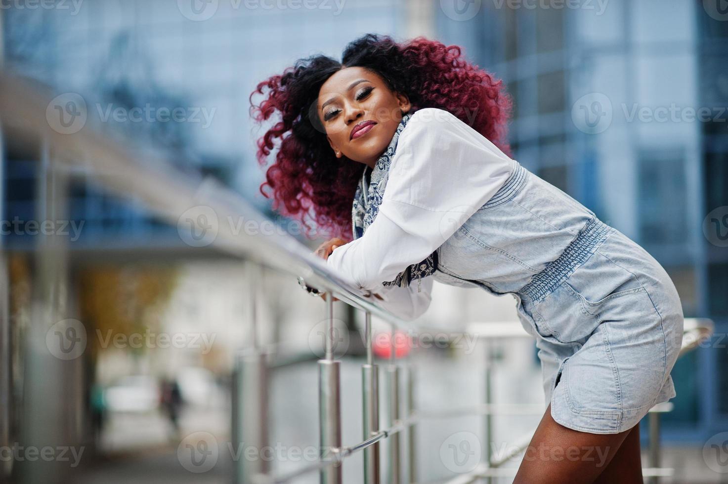Attraktive lockige Afroamerikanerin im Jeanskleid posierte gegen modernes mehrstöckiges Gebäude. foto