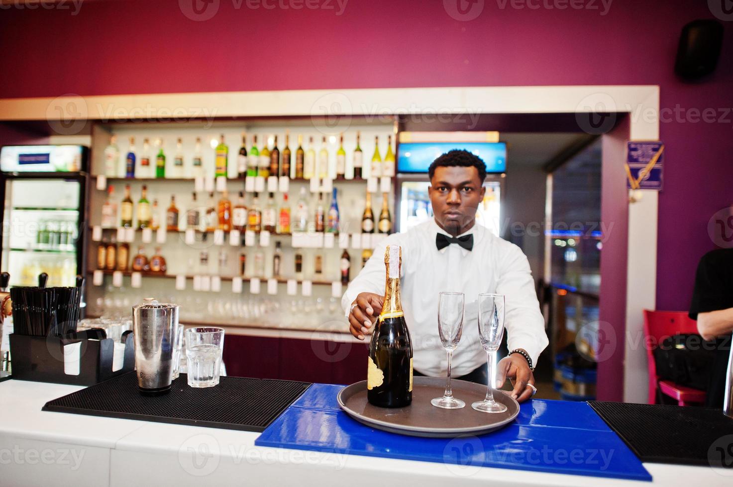 Afroamerikanischer Barkeeper an der Bar mit Champagner und Gläsern auf dem Tablett. foto