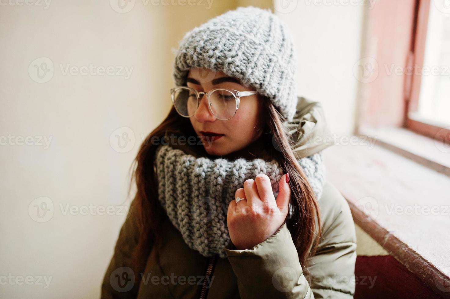 Porträt eines brünetten Mädchens mit grauem Schal und Hut, Brille in der Nähe des Fensters. foto