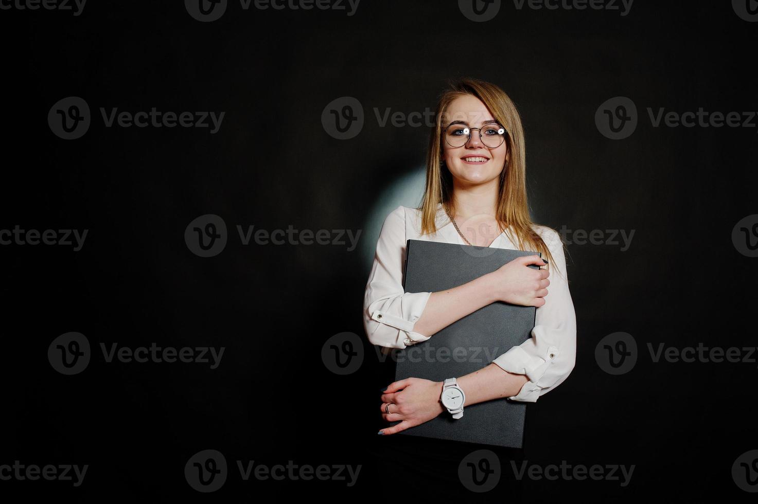 Studioporträt einer blonden Geschäftsfrau mit Brille, weißer Bluse und schwarzem Rock, die einen Laptop vor dunklem Hintergrund hält. erfolgreiche frau und stilvolles mädchenkonzept. foto