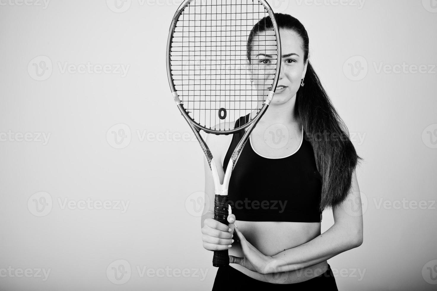 Schwarz-Weiß-Porträt einer schönen jungen Spielerin in Sportkleidung, die einen Tennisschläger hält, während sie vor weißem Hintergrund steht. foto