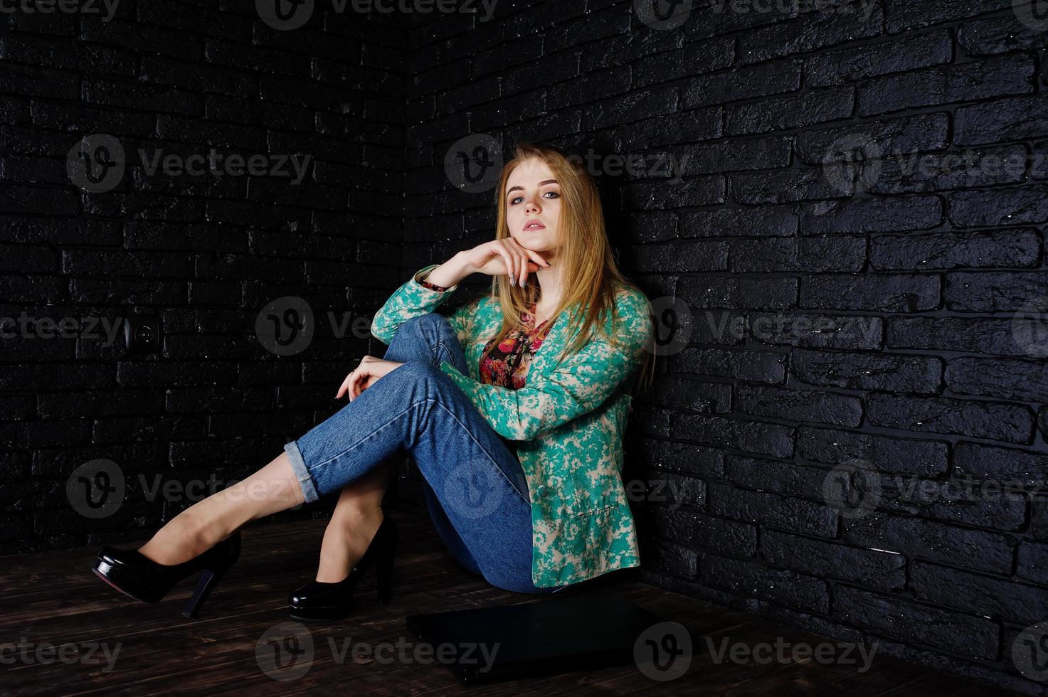 Stilvolles blondes Mädchen in Jacke und Jeans mit Laptop vor schwarzer Ziegelwand im Studio. foto