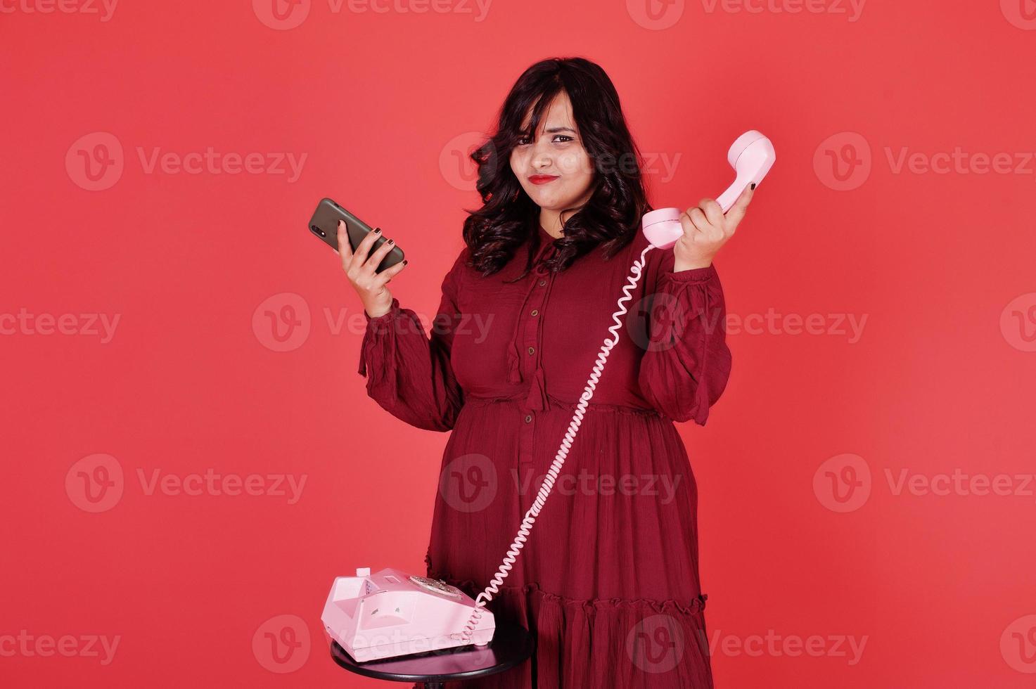 attraktive südasiatische frau in tiefrotem kleiderkleid posierte im studio auf rosa hintergrund und benutzte altes vintage-telefon mit modernem handy. foto