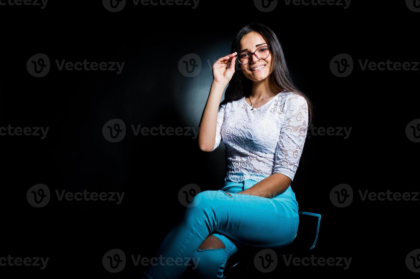 Porträt einer attraktiven jungen Frau in weißem Oberteil und blauer Hose, die mit ihrer Brille im Dunkeln posiert. foto