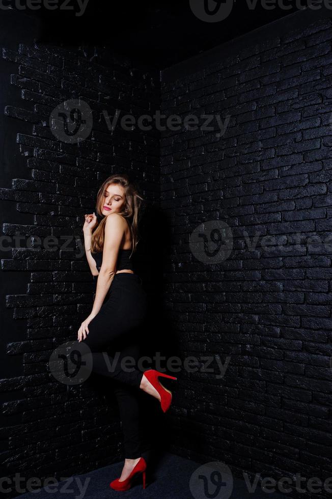 hübsche brünette mädchen tragen schwarze und rote high heels und posieren im studio gegen eine dunkle backsteinmauer. Studio-Model-Porträt. foto