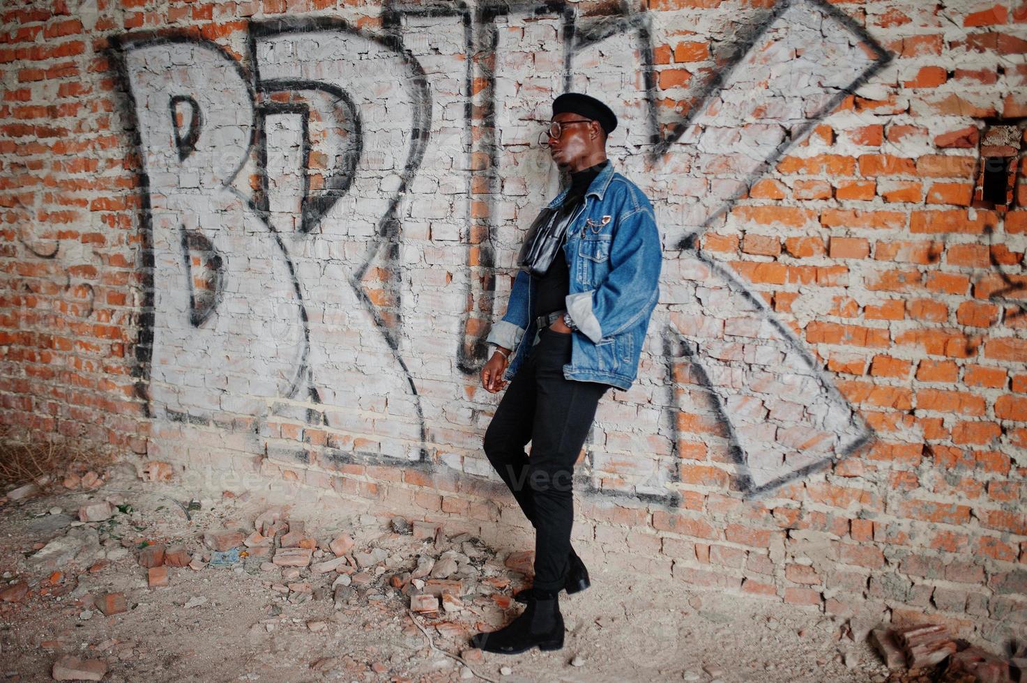 afroamerikanischer mann in jeansjacke, baskenmütze und brille gegen graffiti-ziegelwand mit bruk-schild. foto