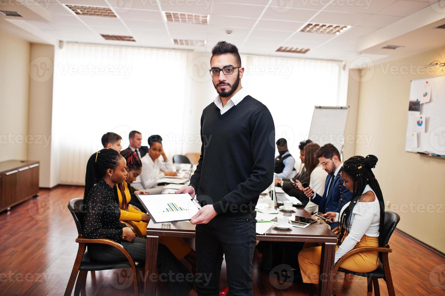 Gesicht eines gutaussehenden arabischen Geschäftsmannes, der eine Zwischenablage auf dem Hintergrund des multirassischen Teamtreffens der Geschäftsleute hält und am Bürotisch sitzt. foto