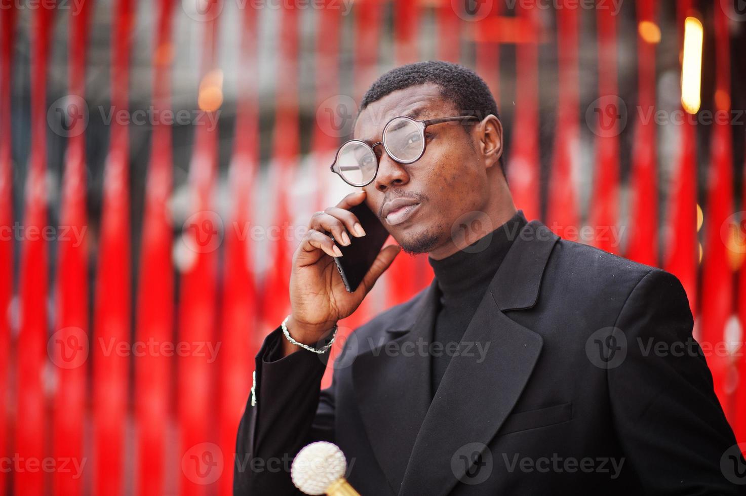 Stilvoller afroamerikanischer Gentleman in eleganter schwarzer Jacke, der einen Retro-Spazierstock als Rohrkolben oder einen Kippstock mit goldenem Diamantkugelgriff hält. reicher modischer afro-mann gegen rote wand. foto