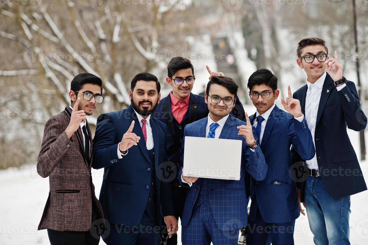 gruppe von sechs indischen geschäftsleuten in anzügen posierte im freien am wintertag in europa, schaute auf laptop und zeigte finger nach oben. foto