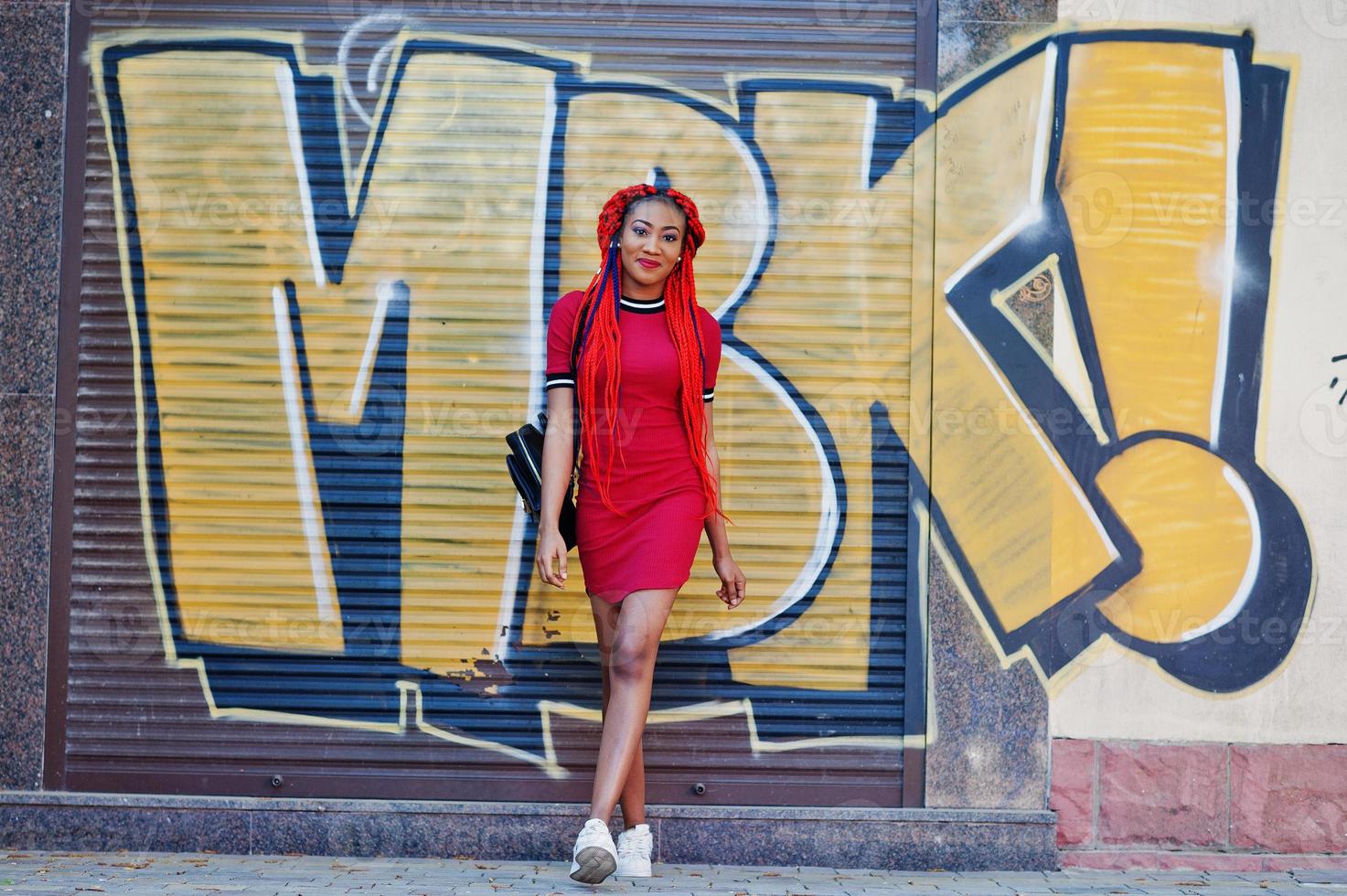 süßes und schlankes afrikanisch-amerikanisches Mädchen in rotem Kleid mit Dreadlocks und Rucksack posierte im Freien auf der Straßenhintergrund-Graffitiwand. stylisches schwarzes Modell. foto