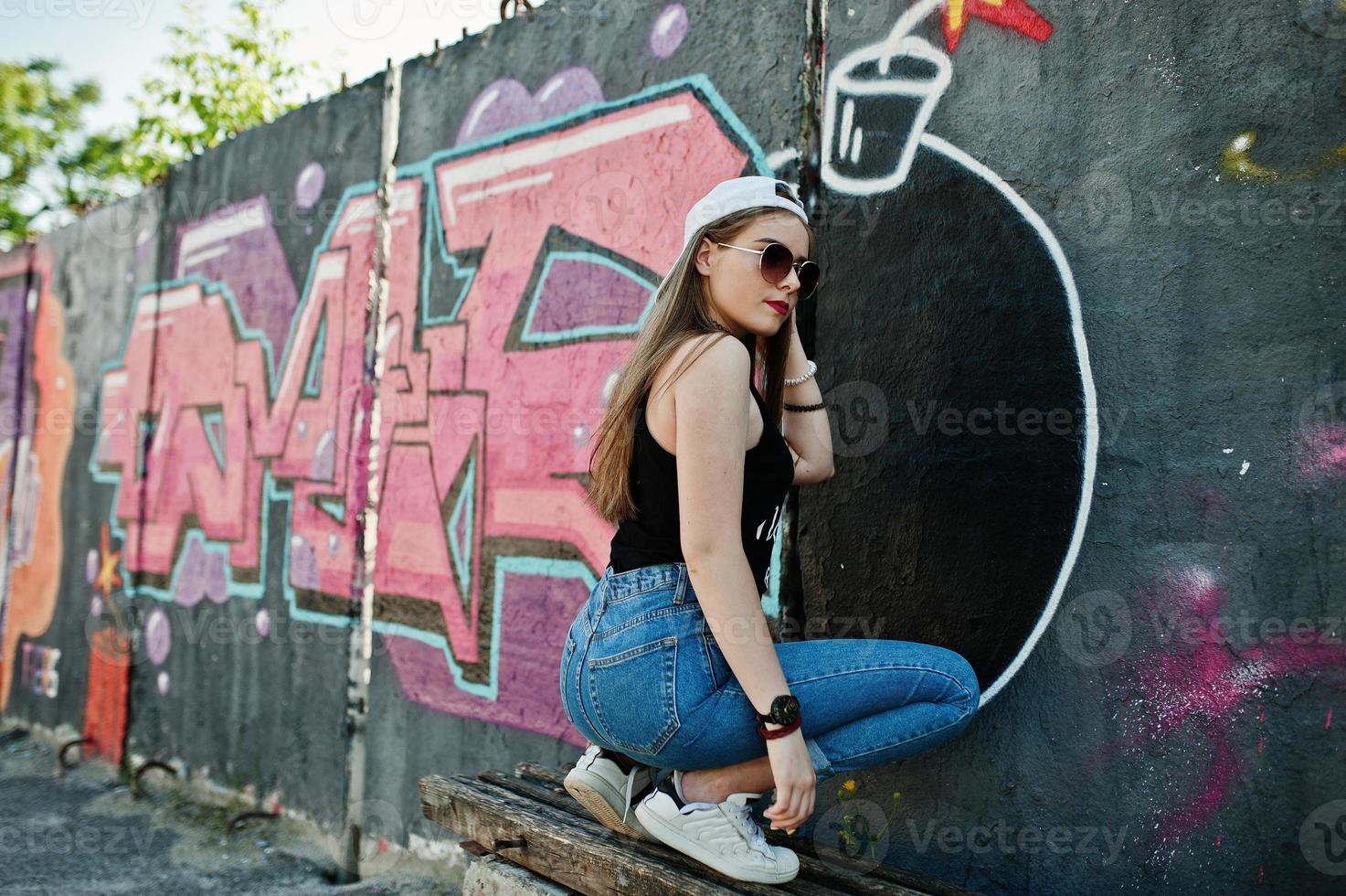Rückseite des Gesäßes stilvolles, lässiges Hipster-Mädchen in Mütze, Sonnenbrille und Jeans, das Musik über Kopfhörer des Mobiltelefons gegen große Graffiti-Wand mit großer TNT-Bombe hört. foto