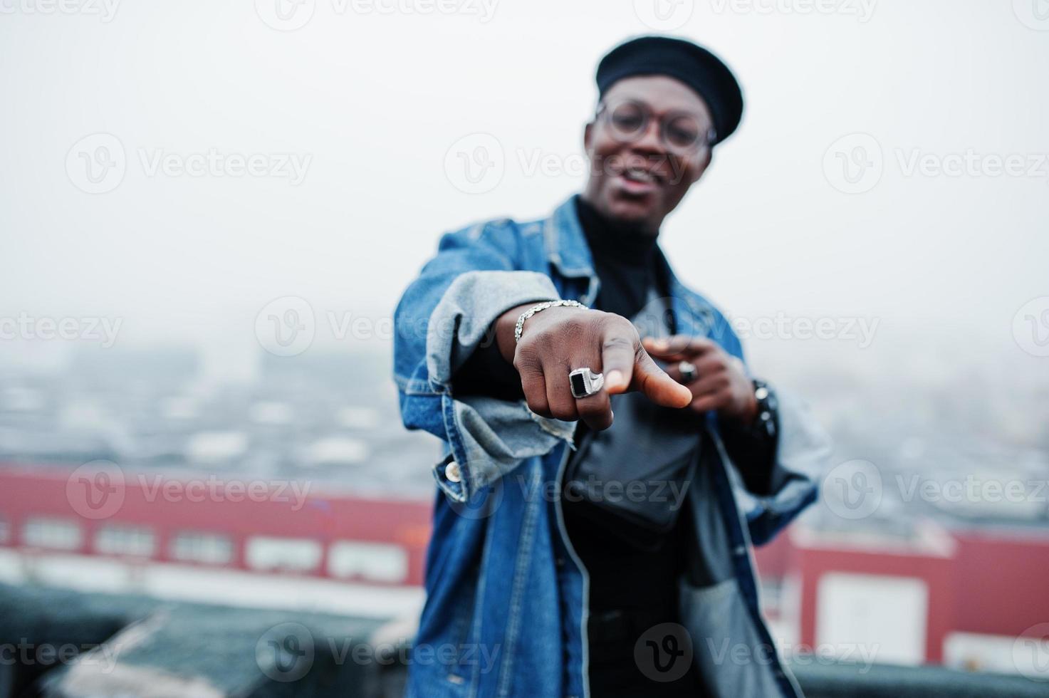 Afroamerikaner in Jeansjacke, Baskenmütze und Brille auf verlassenem Dach zeigt Finger. foto