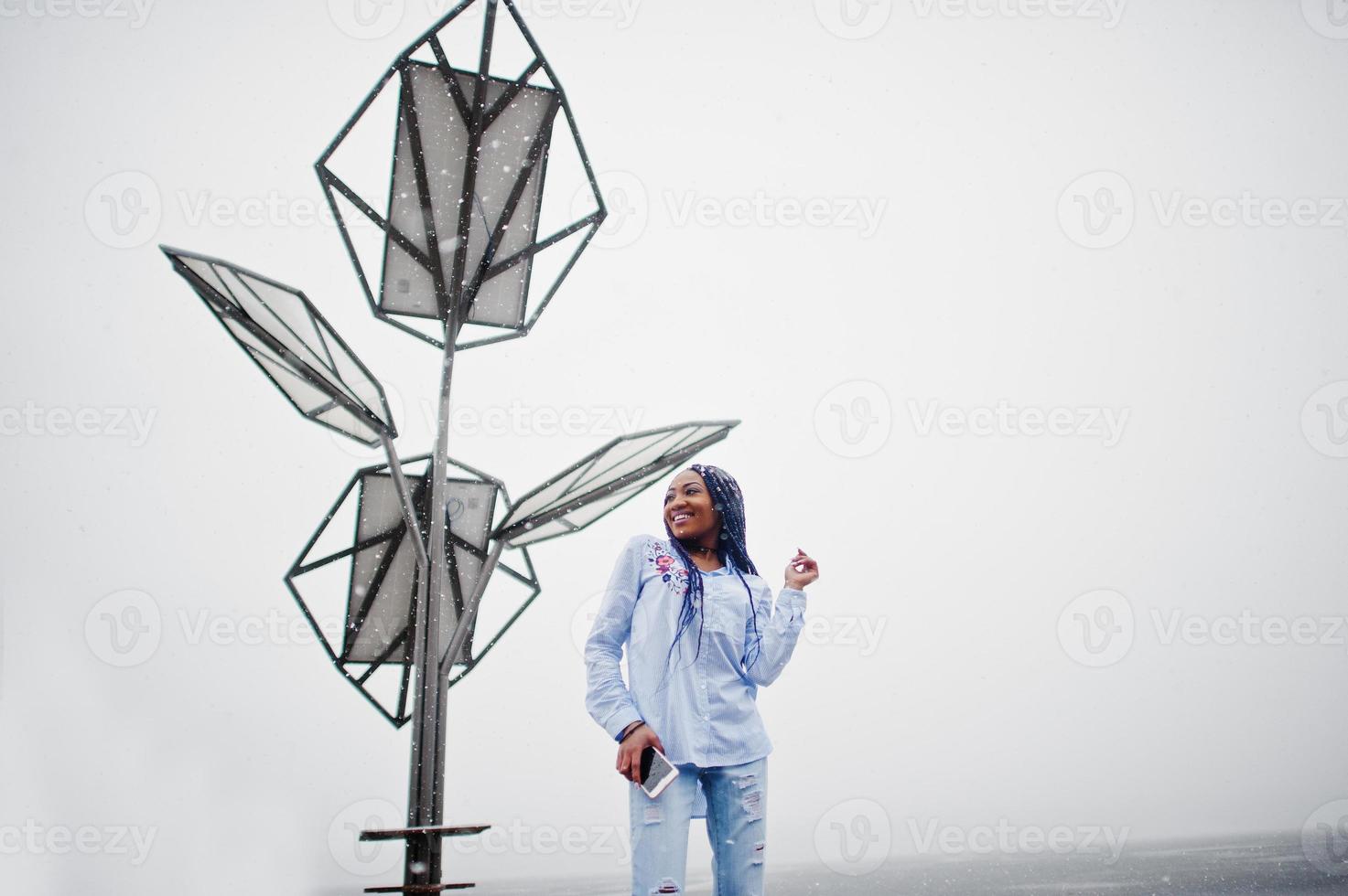 stilvolles afrikanisch-amerikanisches Mädchen mit Dreadlocks, das Handy zur Hand hält, im Freien gegen Solarbatterien bei Schneewetter. foto