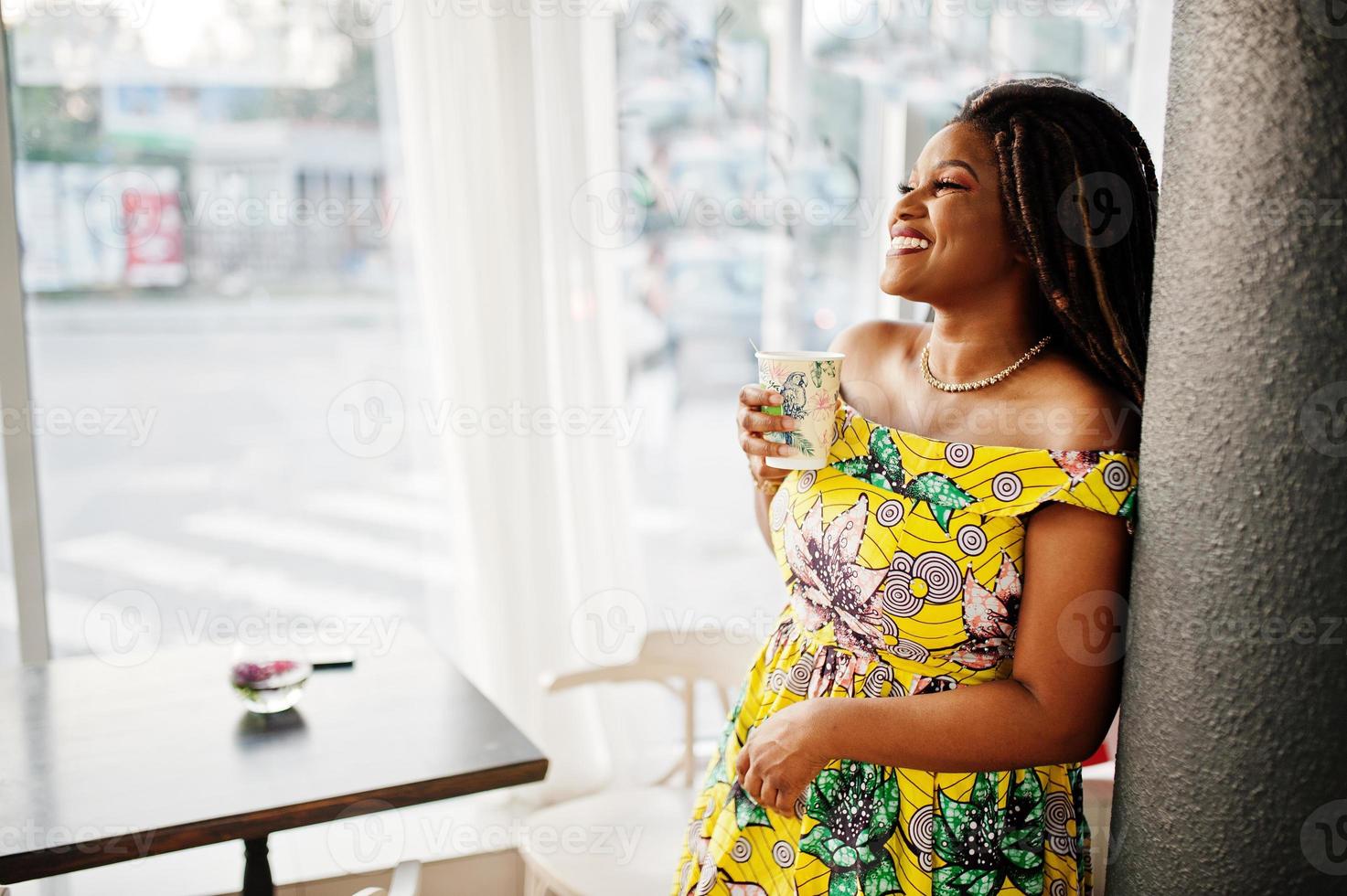 süßes kleines afroamerikanisches mädchen mit dreadlocks, trägt ein farbiges gelbes kleid, posiert im café mit einer tasse kaffee. foto