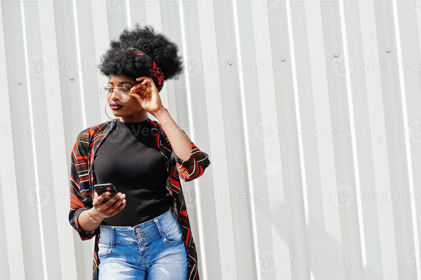 afrikanische frau mit afrohaar, in jeansshorts und brille, die mit handy in der hand gegen eine weiße stahlwand posiert. foto