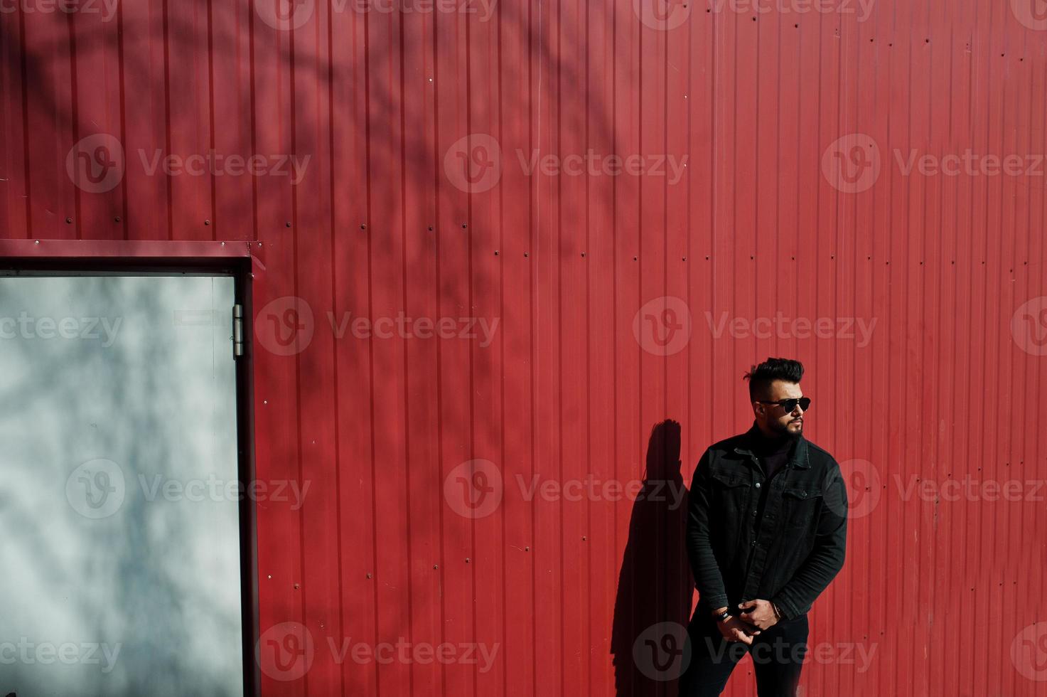 Mode arabischer Mann trägt schwarze Jeansjacke und Sonnenbrille vor rotem Stahlwandhintergrund. stilvoller und modischer arabischer Modelltyp. foto