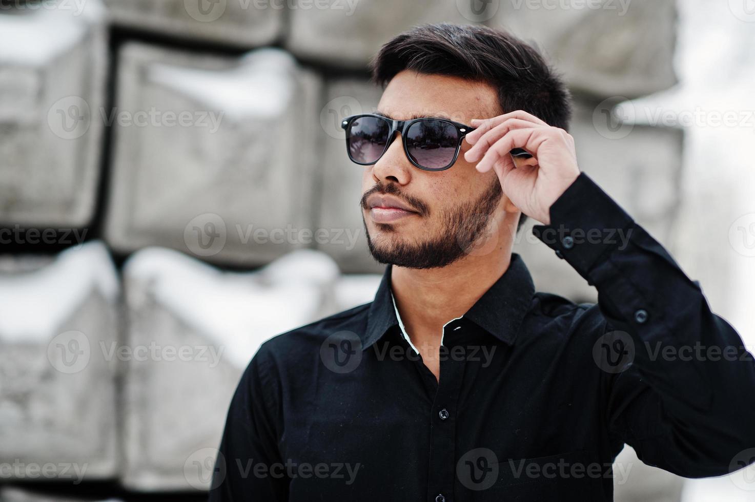 Lässiger junger indischer Mann in schwarzem Hemd und Sonnenbrille posierte gegen Steinblöcke. foto