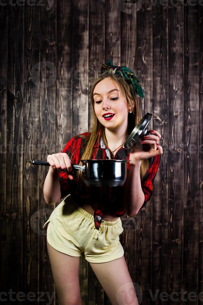 junge lustige hausfrau in kariertem hemd und gelben shorts pin-up-stil mit topf auf hölzernem hintergrund. foto