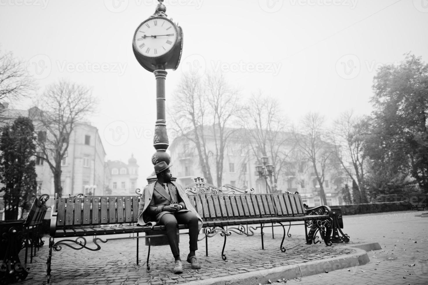 stilvolles afroamerikanisches mannmodell in grauem mantel, jacke, krawatte und rotem hut posierte auf einer bank gegen eine große uhr. Schwarz-Weiß-Foto. foto