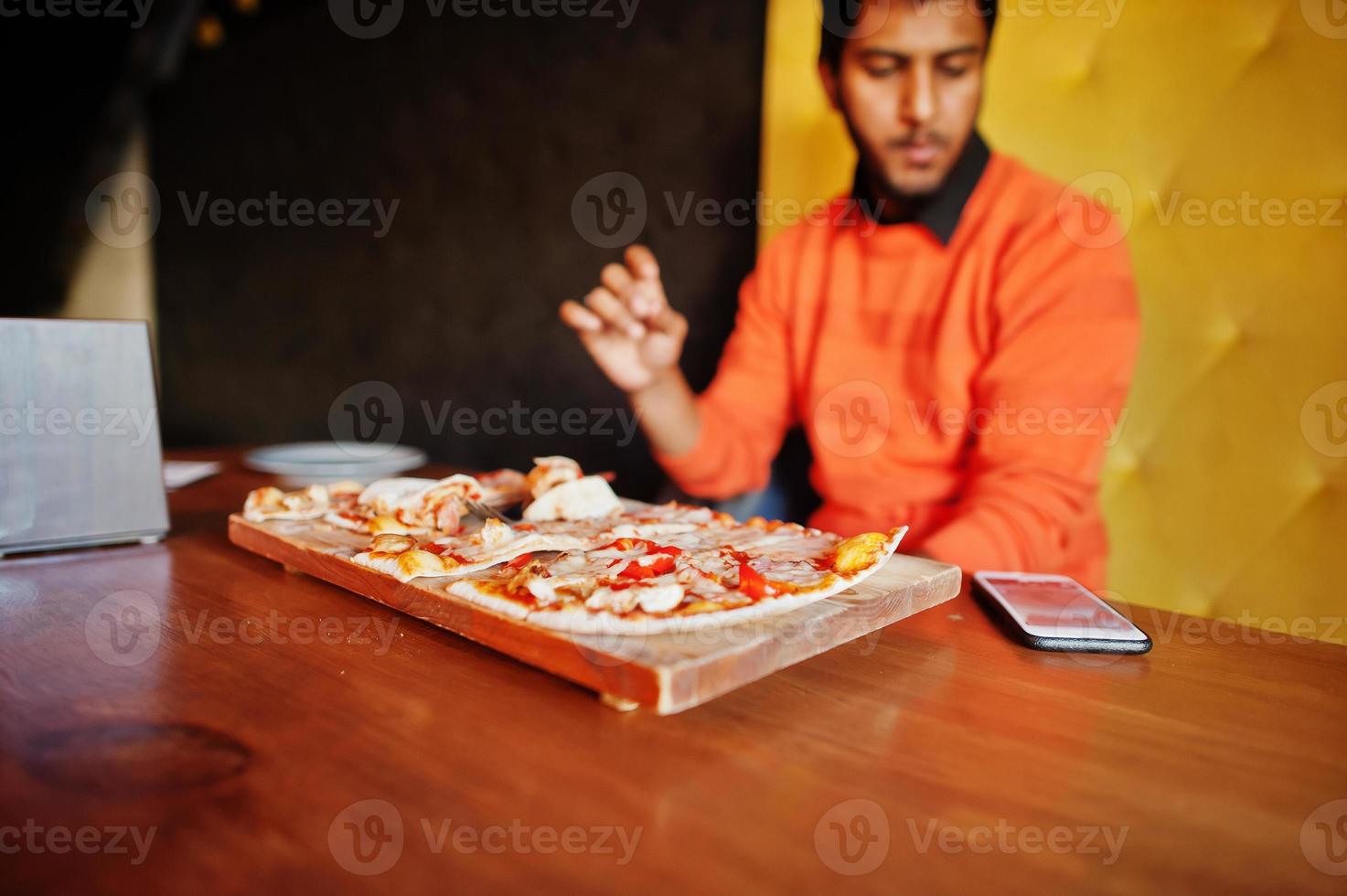 Selbstbewusster junger Inder in orangefarbenem Pullover, der in der Pizzeria sitzt und Pizza isst. foto