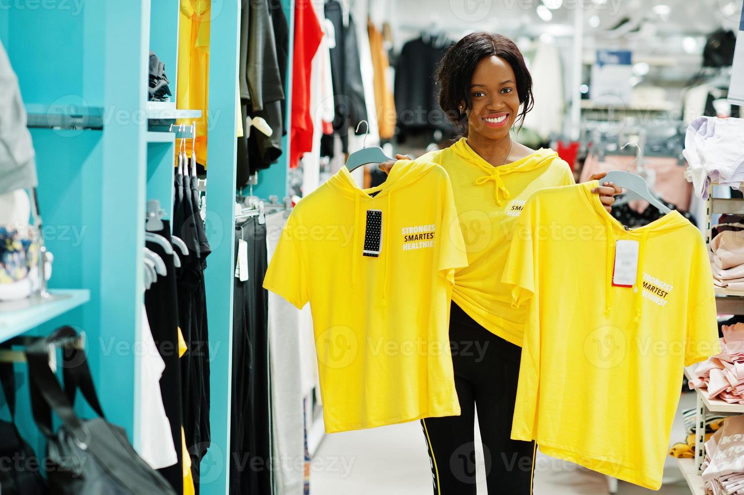 afroamerikanische Frauen in Trainingsanzügen, die im Sportswear-Einkaufszentrum gegen Regale einkaufen. sie wählt gelbes t-shirt. Sportgeschäft Thema. foto