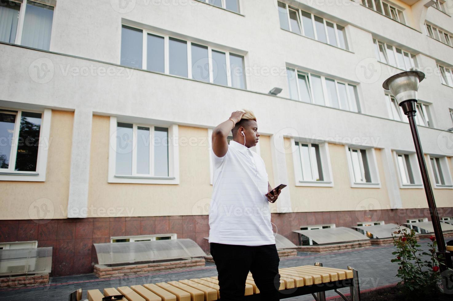 stilvoller arabischer muslimischer junge mit ursprünglich haaren, der auf straßen posiert, auf einer bank sitzt und musik über kopfhörer des telefons hört. foto