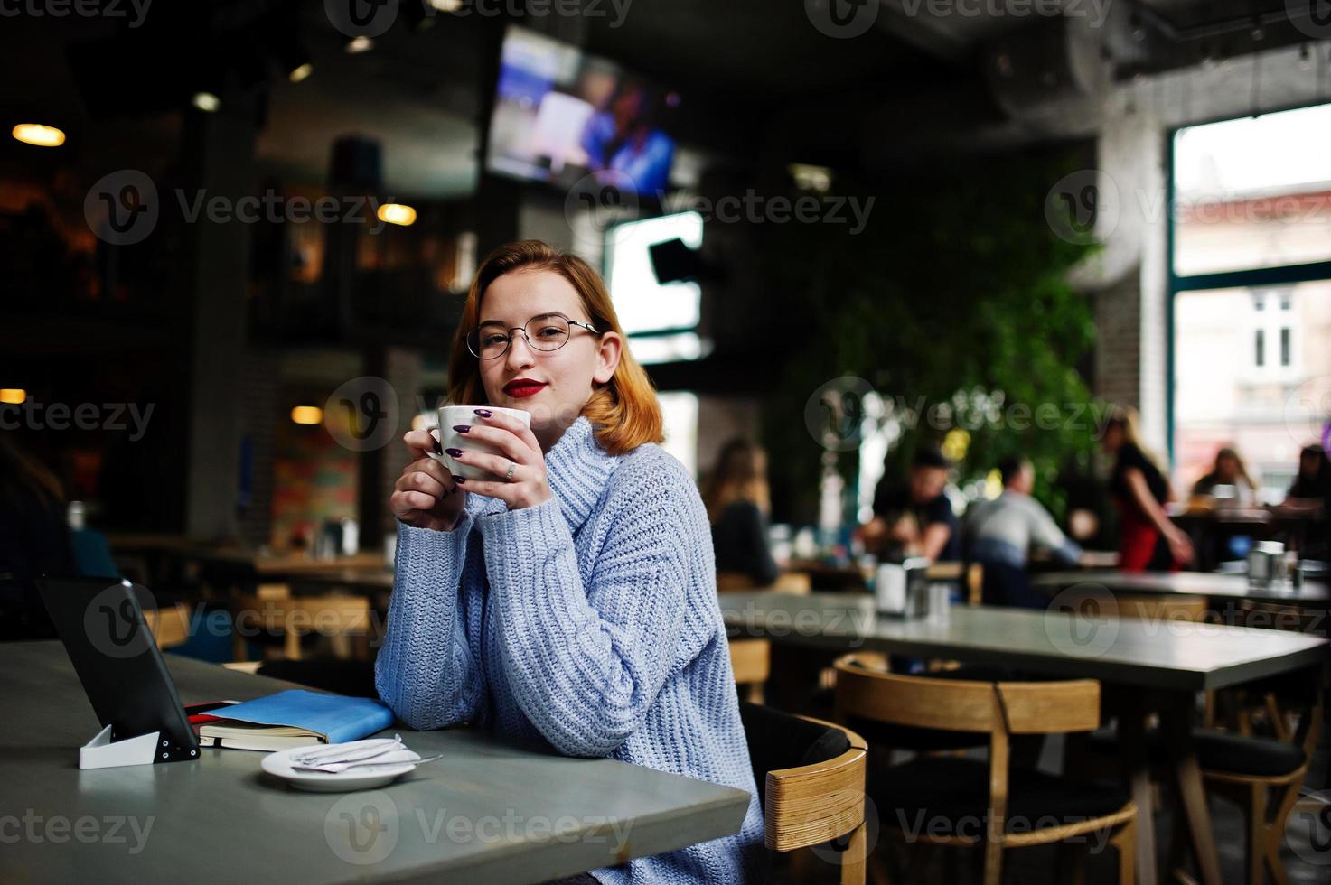 Fröhliche junge schöne rothaarige Frau mit Brille, die ihr Telefon, Touchpad und Notebook benutzt, während sie an ihrem Arbeitsplatz im Café mit einer Tasse Kaffee sitzt. foto
