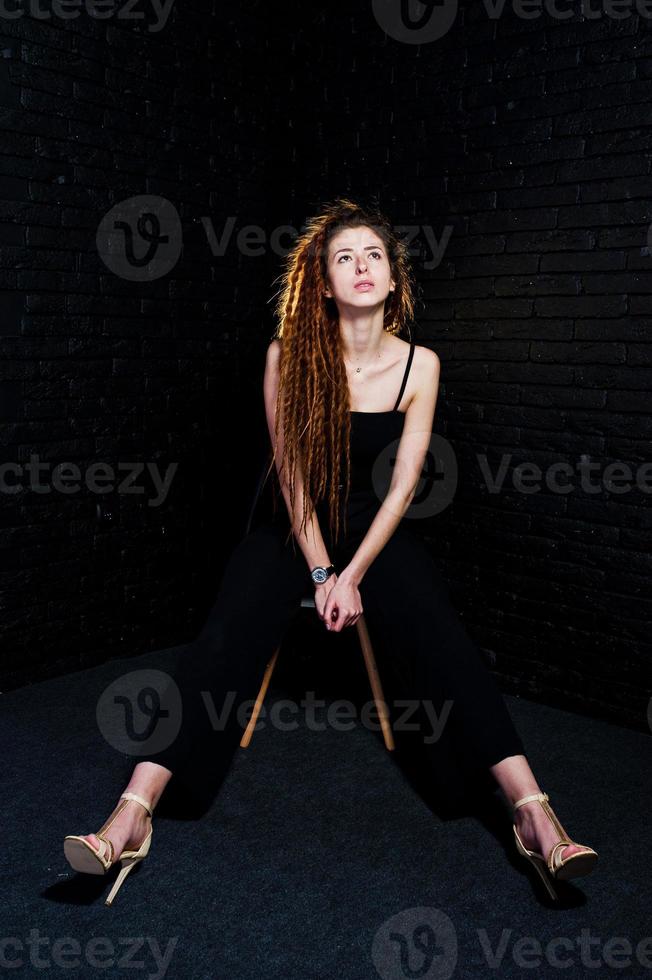 Studioaufnahme eines Mädchens in Schwarz auf einem Stuhl mit Dreadlocks auf Backsteinhintergrund. foto