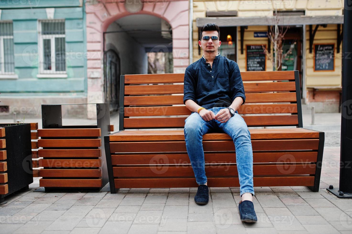 stylischer indischer modellmann in lässiger kleidung, schwarzem hemd und sonnenbrille posierte im freien auf der straße von indien und saß auf einer bank. foto