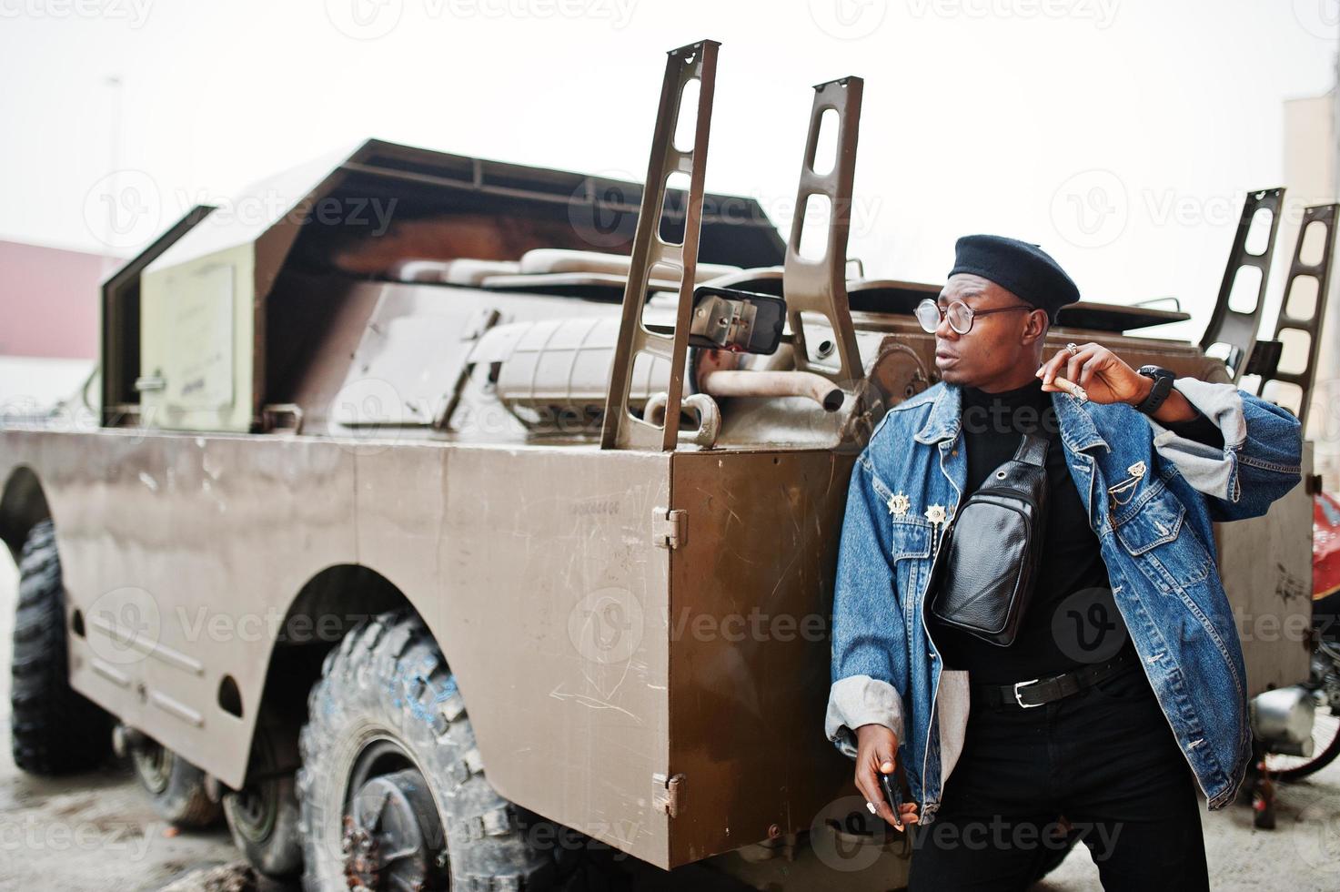 afroamerikanischer mann in jeansjacke, baskenmütze und brille, rauchende zigarre und posierte gegen btr militärisches gepanzertes fahrzeug. foto