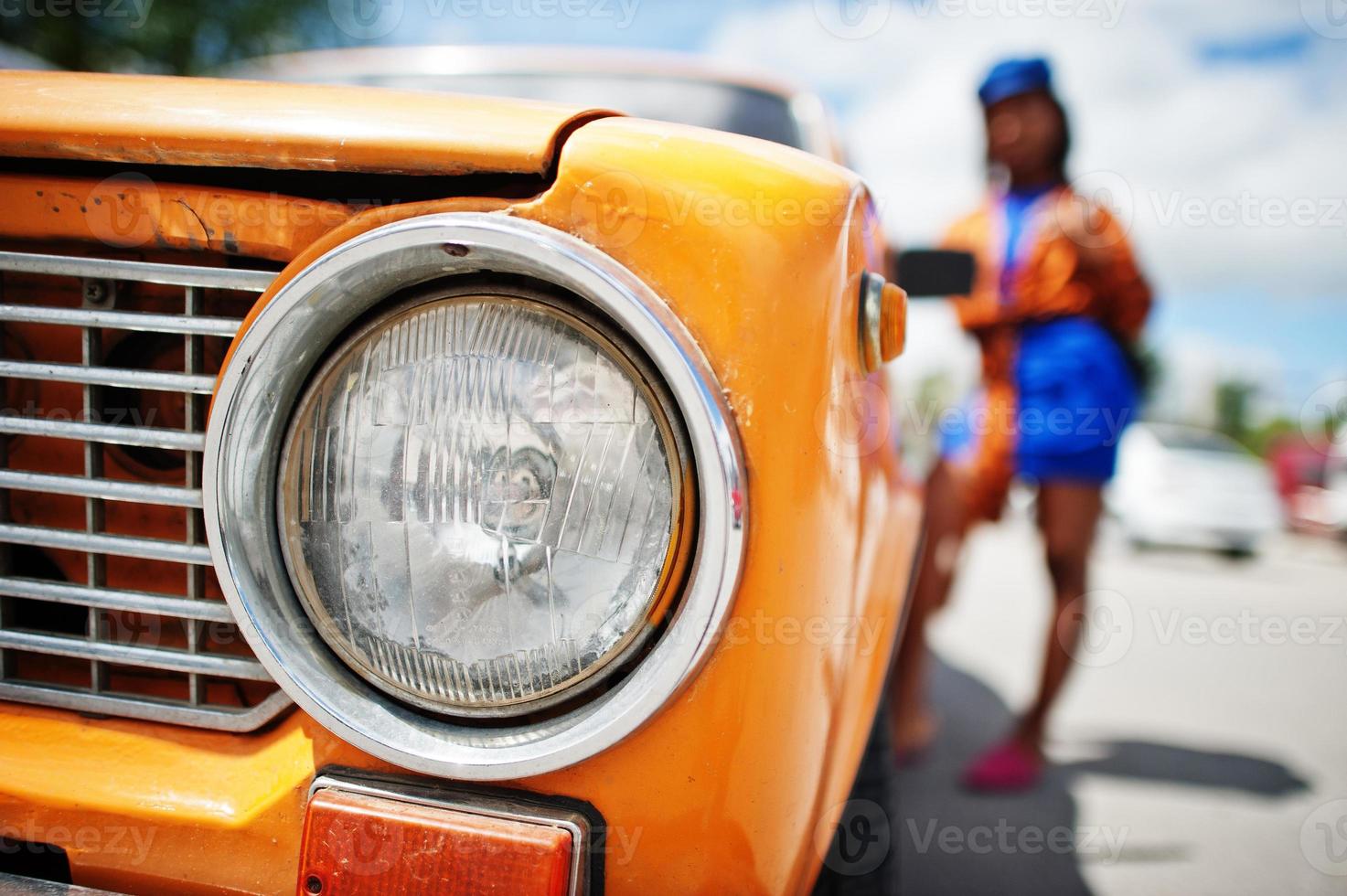 Schöne afrikanisch-amerikanische Dame mit Sonnenbrille, die in der Nähe eines orangefarbenen klassischen Retro-Autos steht. foto