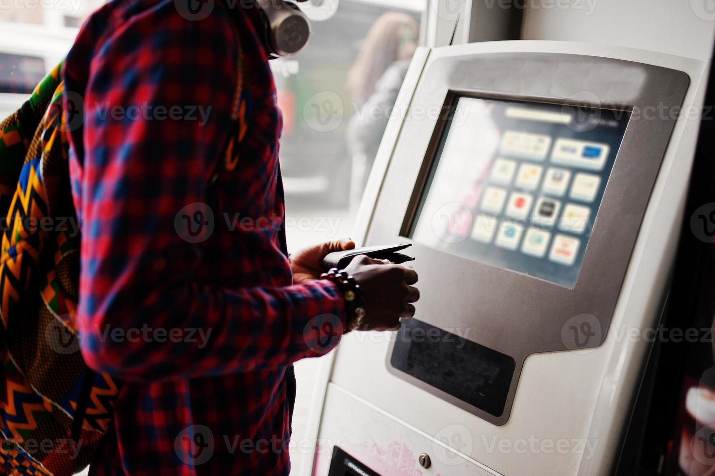 afroamerikanischer mann im karierten hemd mit rucksack, der geldbörse in der hand gegen atm hält. Reisender des schwarzen Mannes. foto