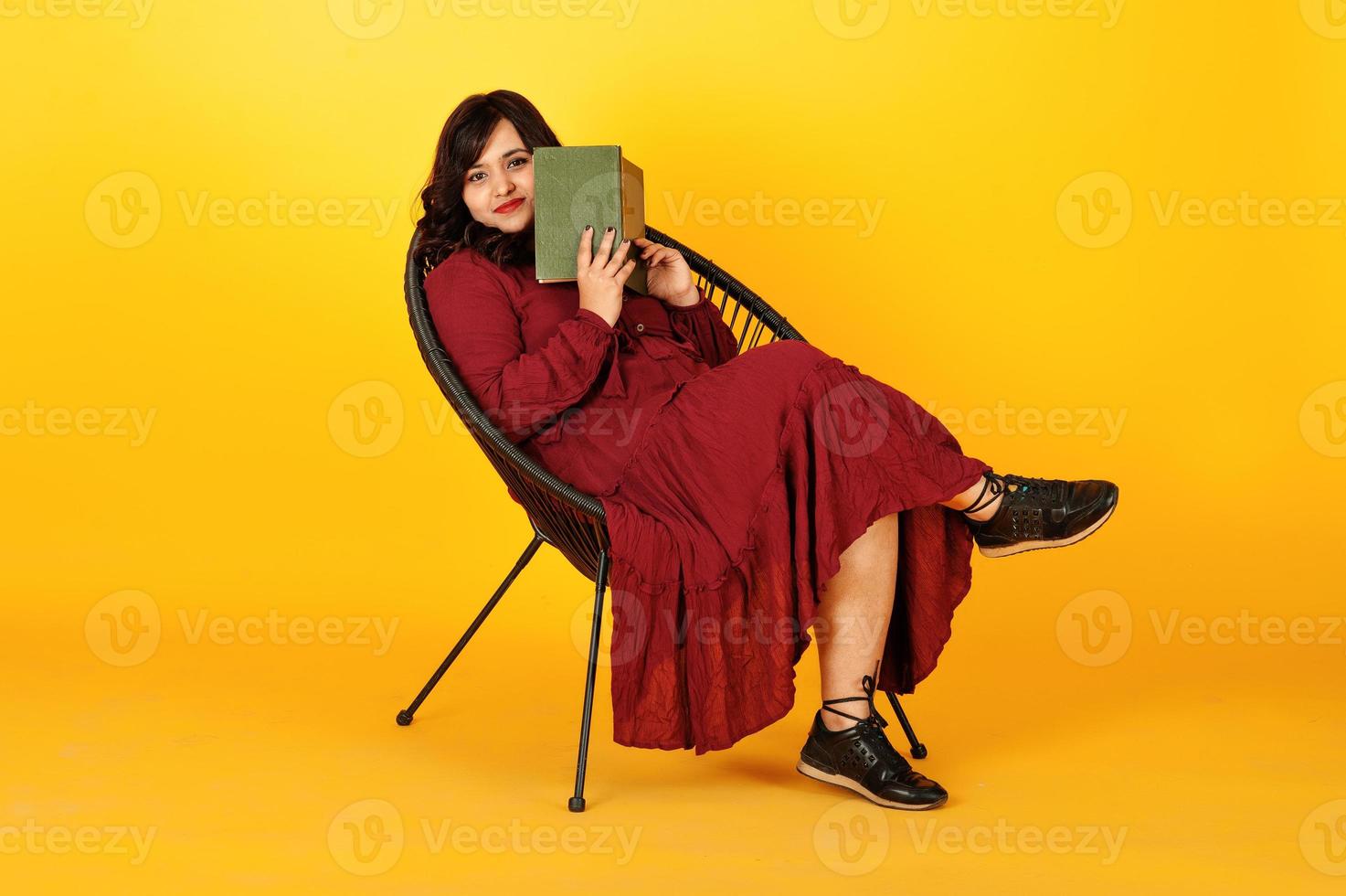 attraktive südasiatische frau in tiefrotem kleiderkleid posierte im studio auf gelbem hintergrund sitzend auf stuhl mit buch. foto