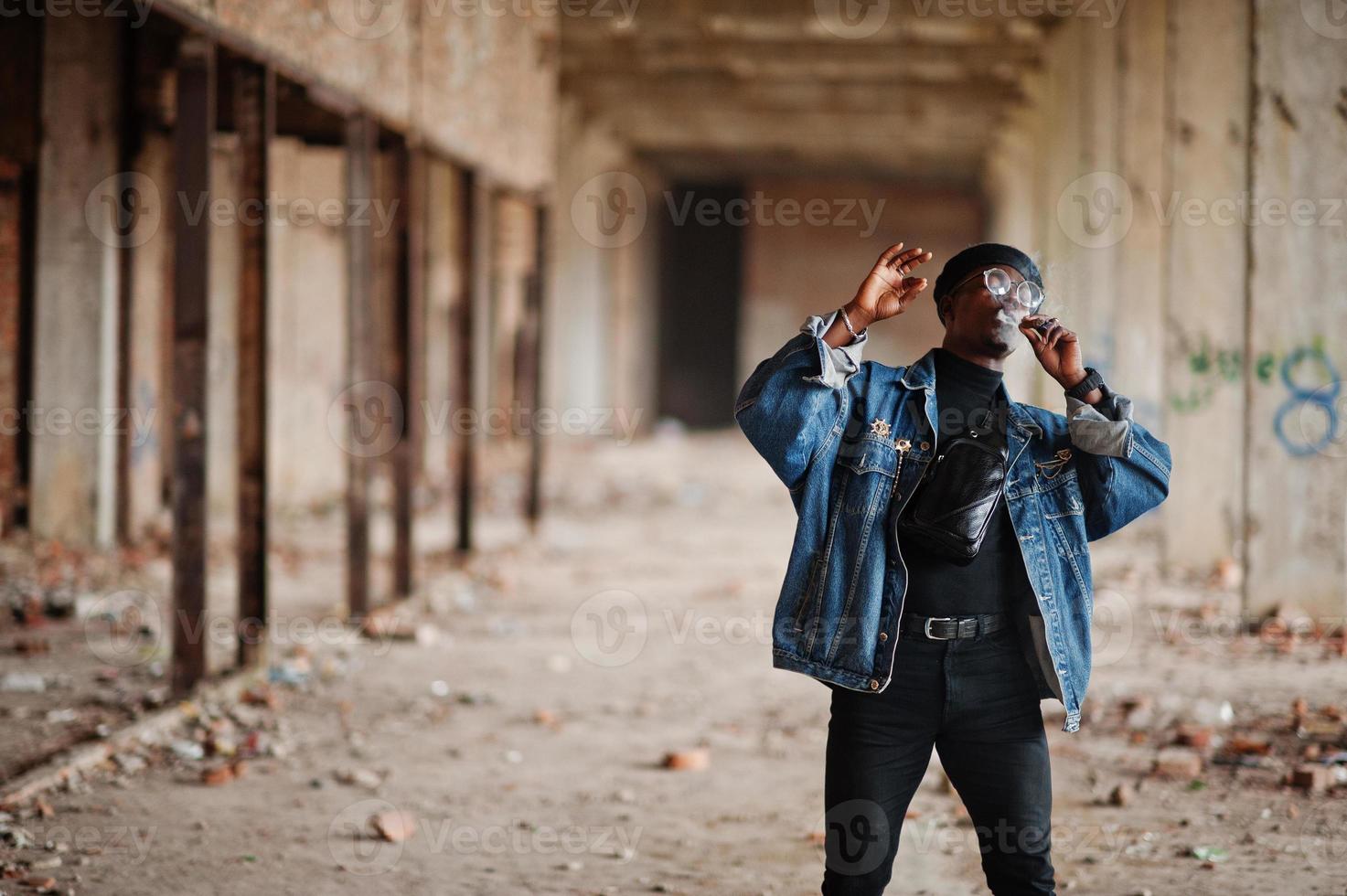 afroamerikanischer mann in jeansjacke, baskenmütze und brille, rauchende zigarre in verlassener fabrik. foto