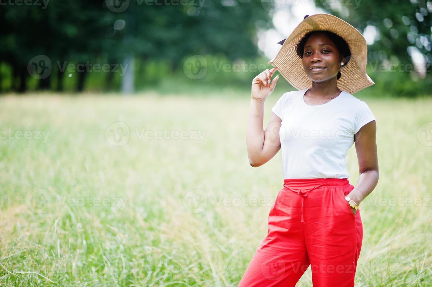 Porträt einer wunderschönen afroamerikanischen Frau der 20er Jahre mit Sommerhut, roten Hosen und weißem T-Shirt, die auf grünem Gras im Park posiert. foto