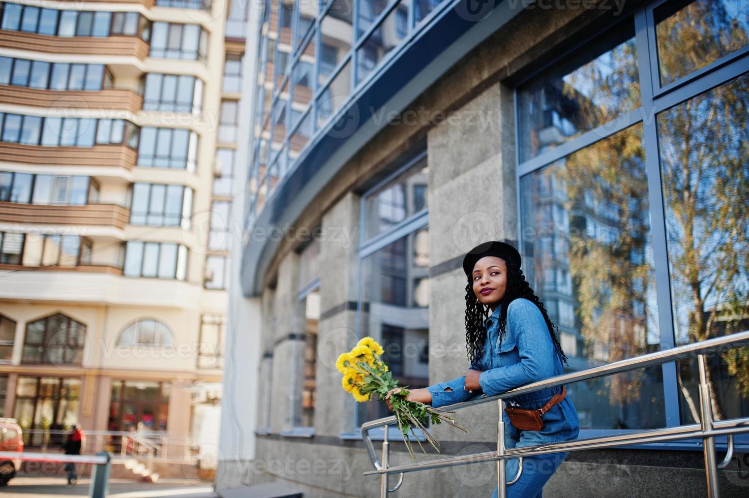 Stilvolle, modische afroamerikanische Frauen in Jeans und schwarzer Baskenmütze mit gelbem Blumenstrauß posierten im Freien an sonnigen Tagen vor blauem, modernem Gebäude. foto