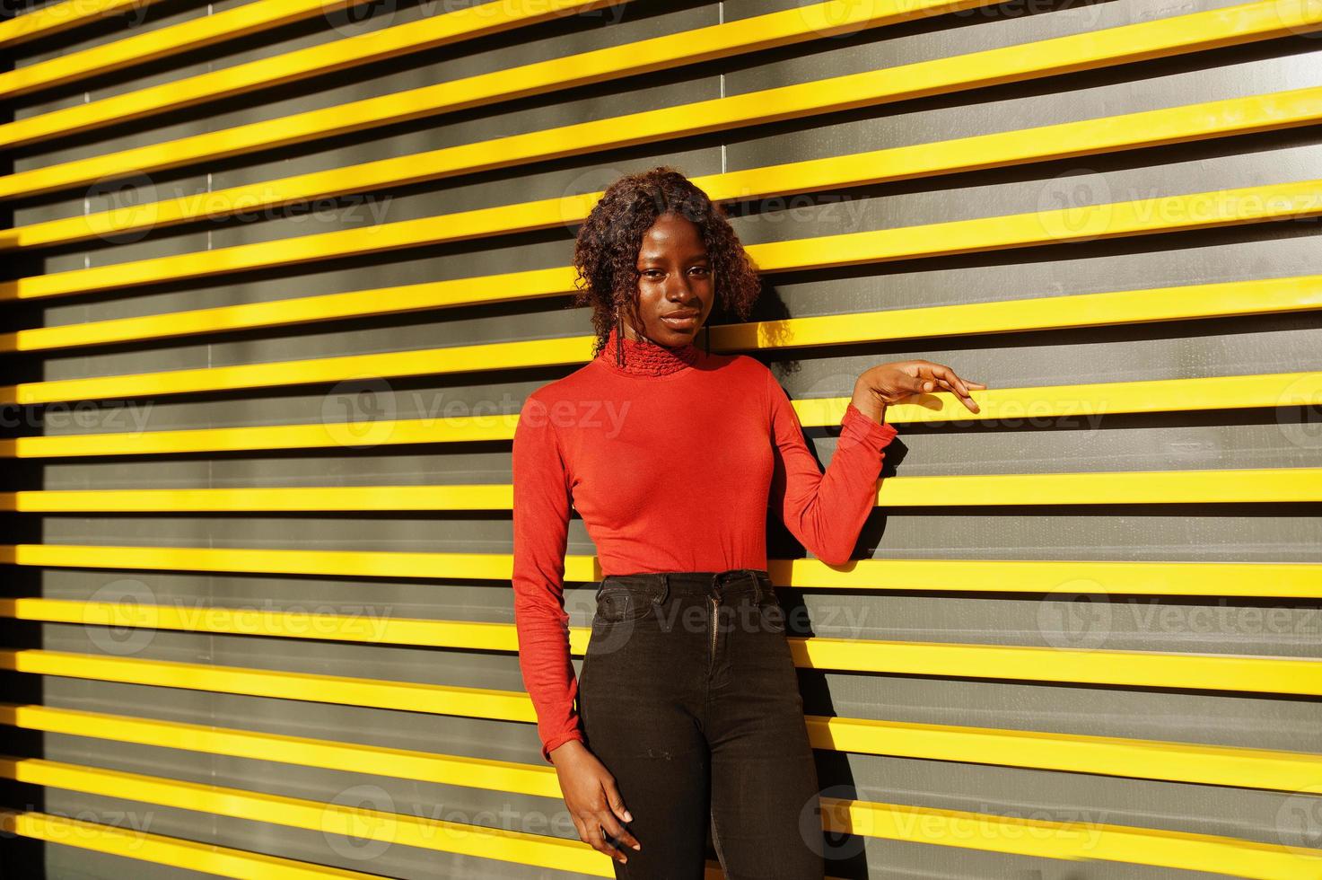 Porträt einer lockigen Afrikanerin mit modischem rotem Rollkragenpullover, die an der gelben Wand mit modernen Linien posiert. foto