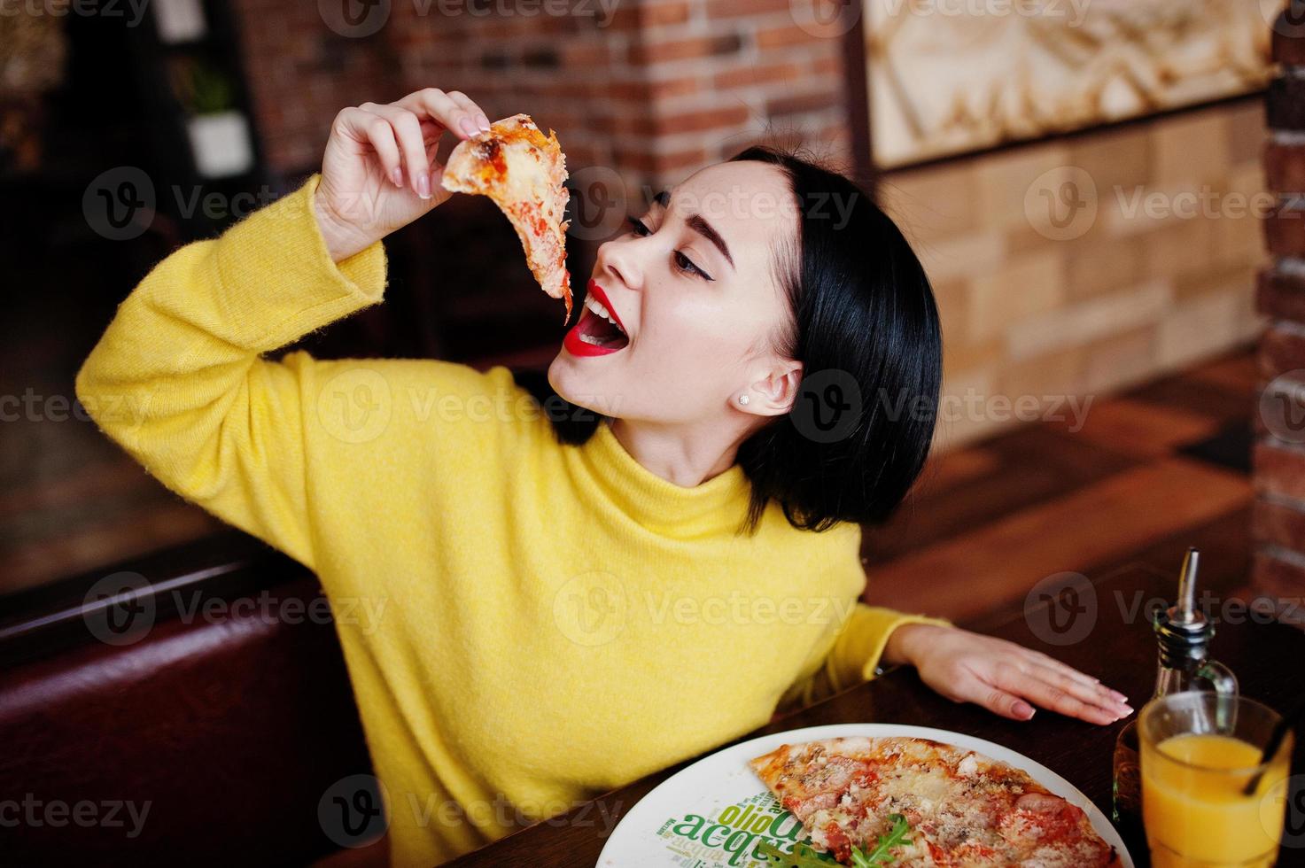 lustiges brünettes mädchen im gelben pullover, der pizza im restaurant isst. foto