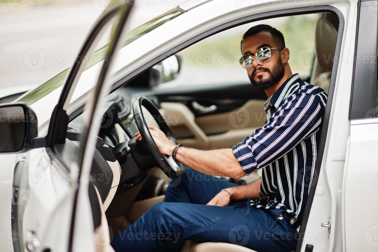 erfolgreicher arabischer mann in gestreiftem hemd und sonnenbrille posiert hinter dem lenkrad seines weißen suv-autos. stilvolle arabische männer im transport. foto
