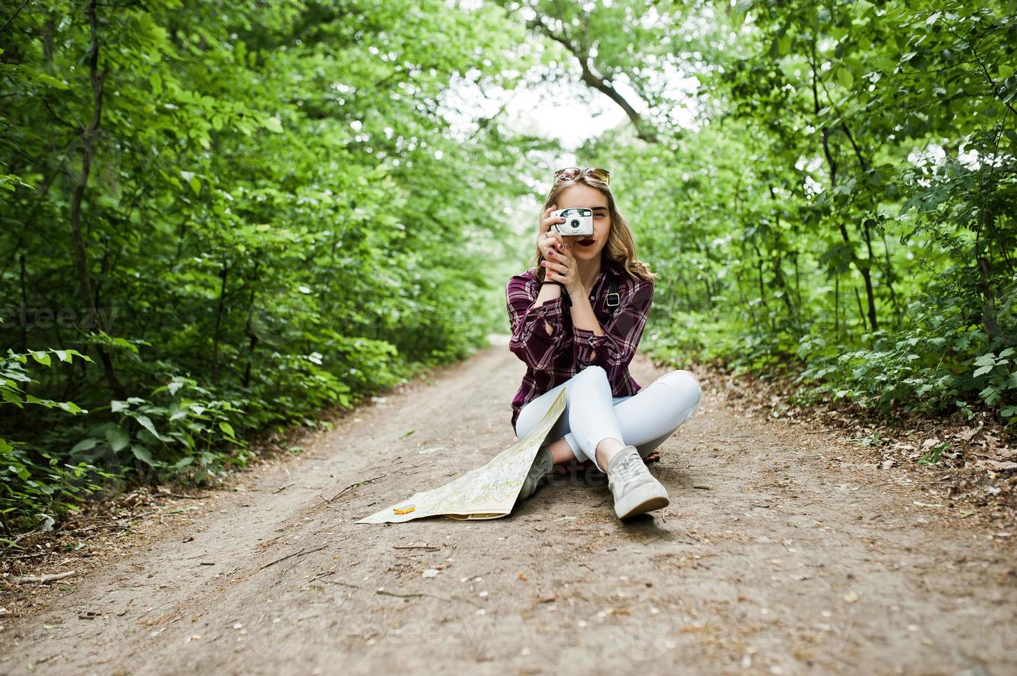 Porträt eines attraktiven blonden Mädchens mit einer Karte, das im Wald sitzt und Fotos macht.