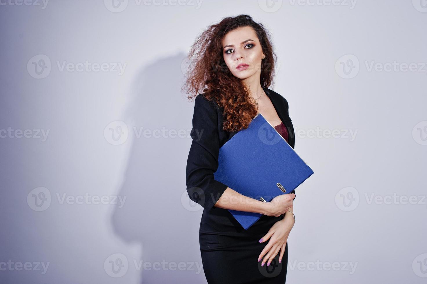 brünettes lockiges Mädchen in schwarzer Bürojacke mit Rock, auf BH mit Ordner mit Dokumenten in Händen auf Studio vor weißem Hintergrund. sexy Geschäftsfrau. foto