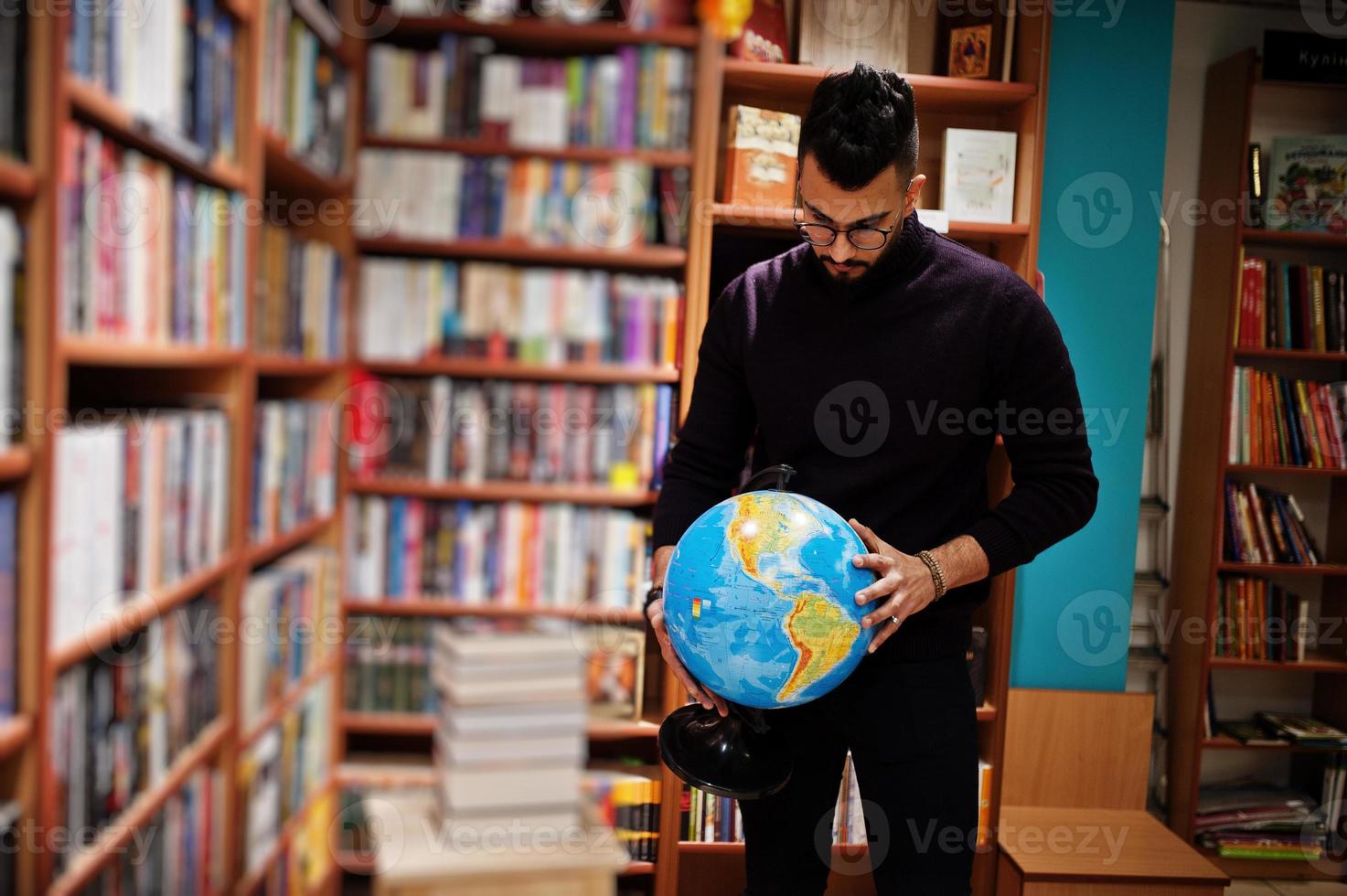 großer, intelligenter arabischer student, trägt einen violetten rollkragenpullover und eine brille, in der bibliothek, die erdkugel an den händen hält. foto