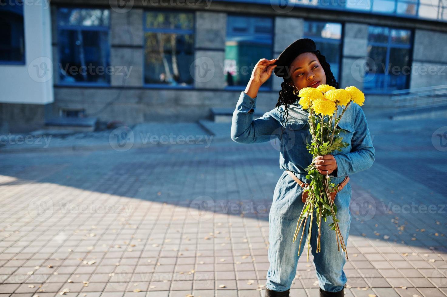 Stilvolle, modische afroamerikanische Frauen in Jeans und schwarzer Baskenmütze mit gelbem Blumenstrauß posierten im Freien an sonnigen Tagen vor blauem, modernem Gebäude. foto