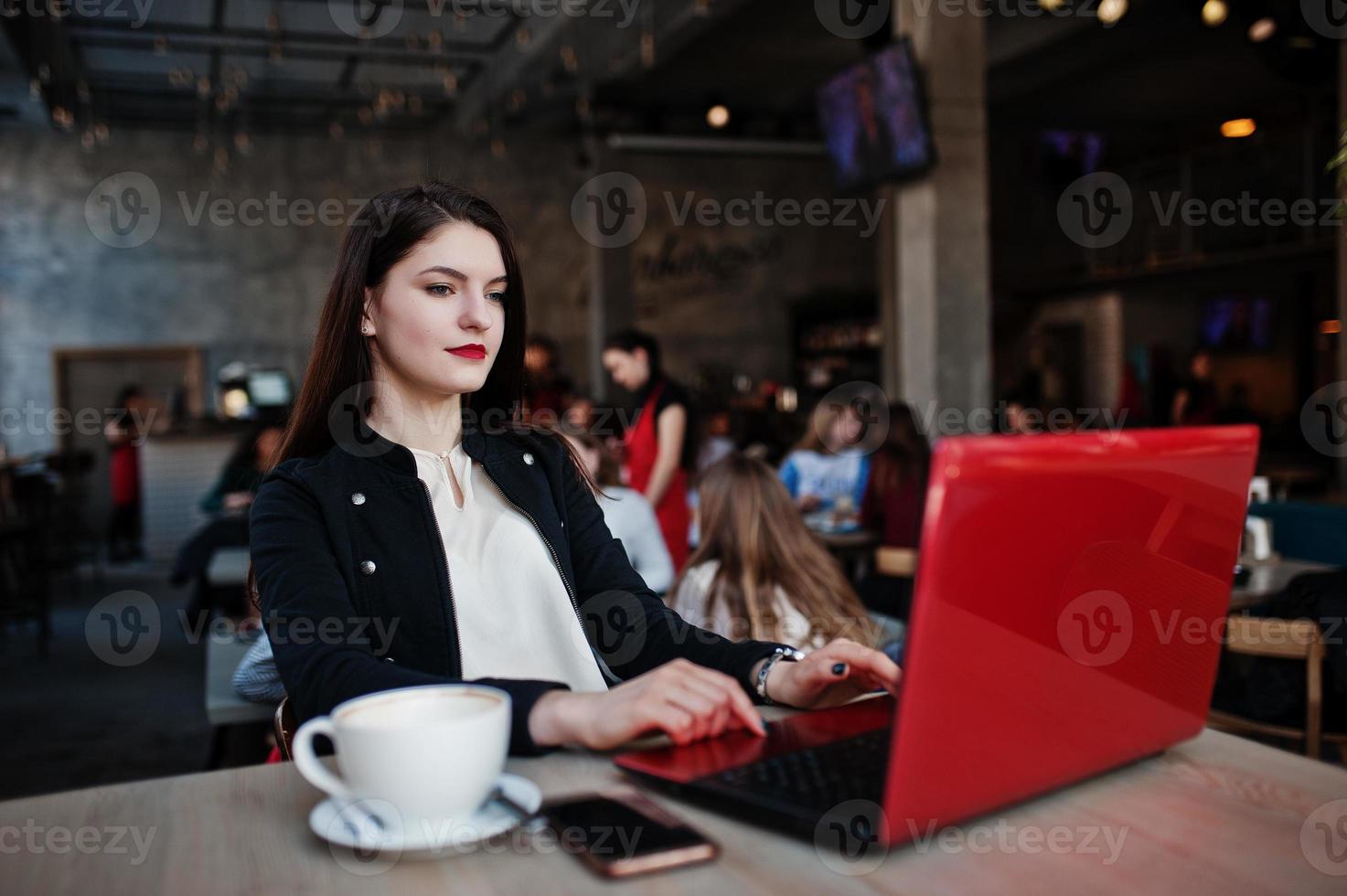 Brünettes Mädchen sitzt im Café mit einer Tasse Cappuccino und arbeitet mit einem roten Laptop. foto